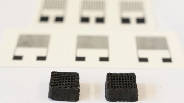 Dva 3D vytlačené kvádre z biologicky odbúrateľného e-atramentu (každý meria 1 cm na šírku). V pozadí sú vidieť vytlačené elektronické senzory.